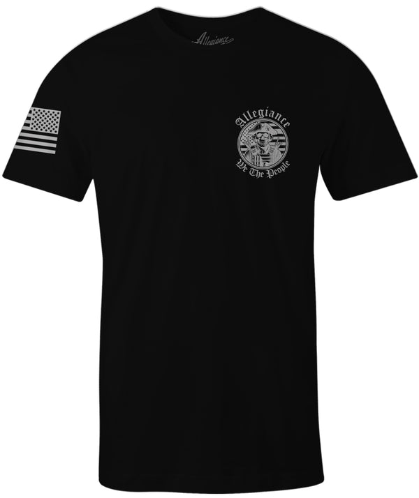 Black t-shirt with small chest print of a skull patriot wearing a tricorn hat and tactical vest, surrounded by the text 'Allegiance We The People'; right sleeve displays a forward-facing American flag